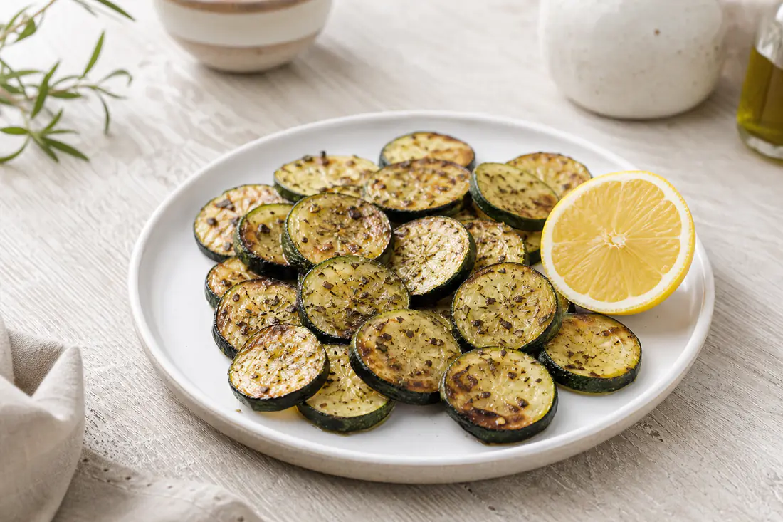 Rodelas de courgette douradas nas bordas, temperadas com orégãos e pimenta, num prato branco com um limão partido ao lado