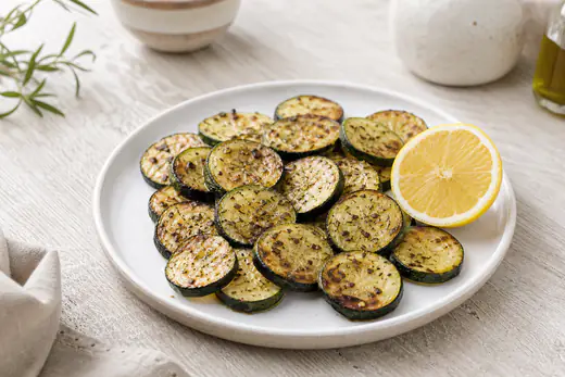 Rodelas de courgette douradas nas bordas, temperadas com orégãos e pimenta, num prato branco com um limão partido ao lado