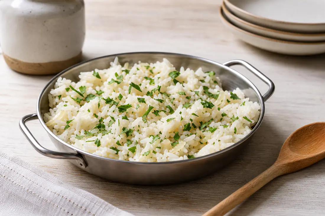 Arroz branco solto numa forma redonda de metal, com cebola e salsa picada por cima, ao lado de uma colher de pau