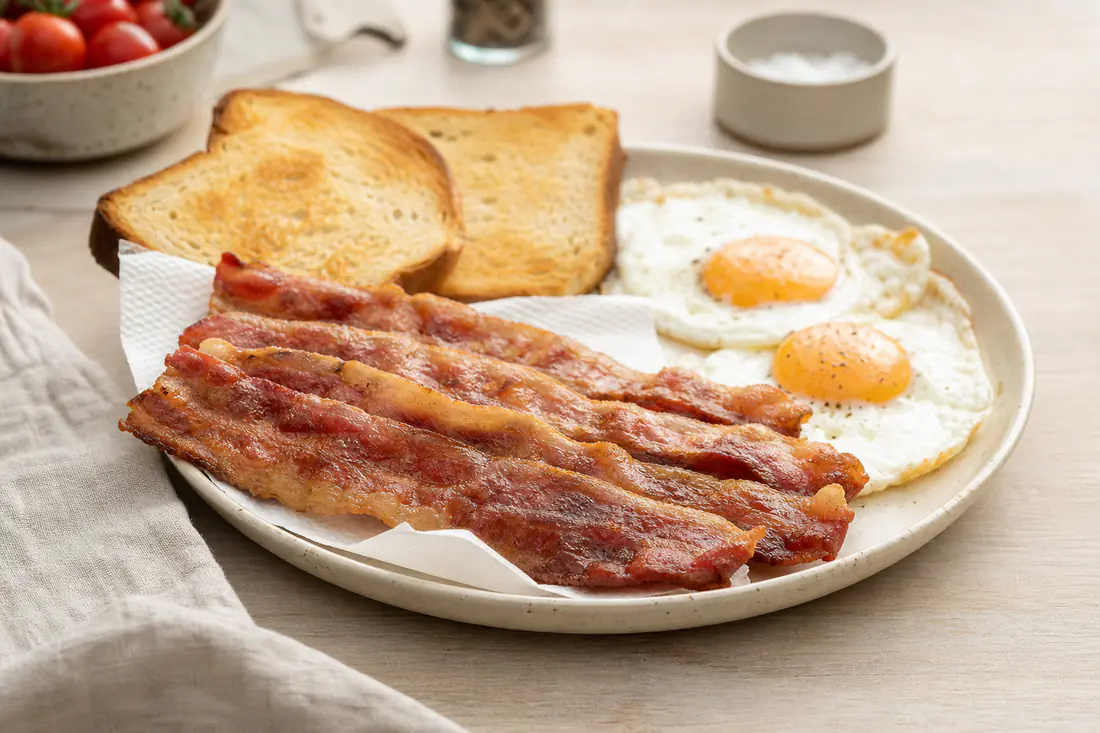 Fatias de bacon crocante e douradas, bem esticadas, numa travessa com papel de cozinha, ao lado de ovos estrelados