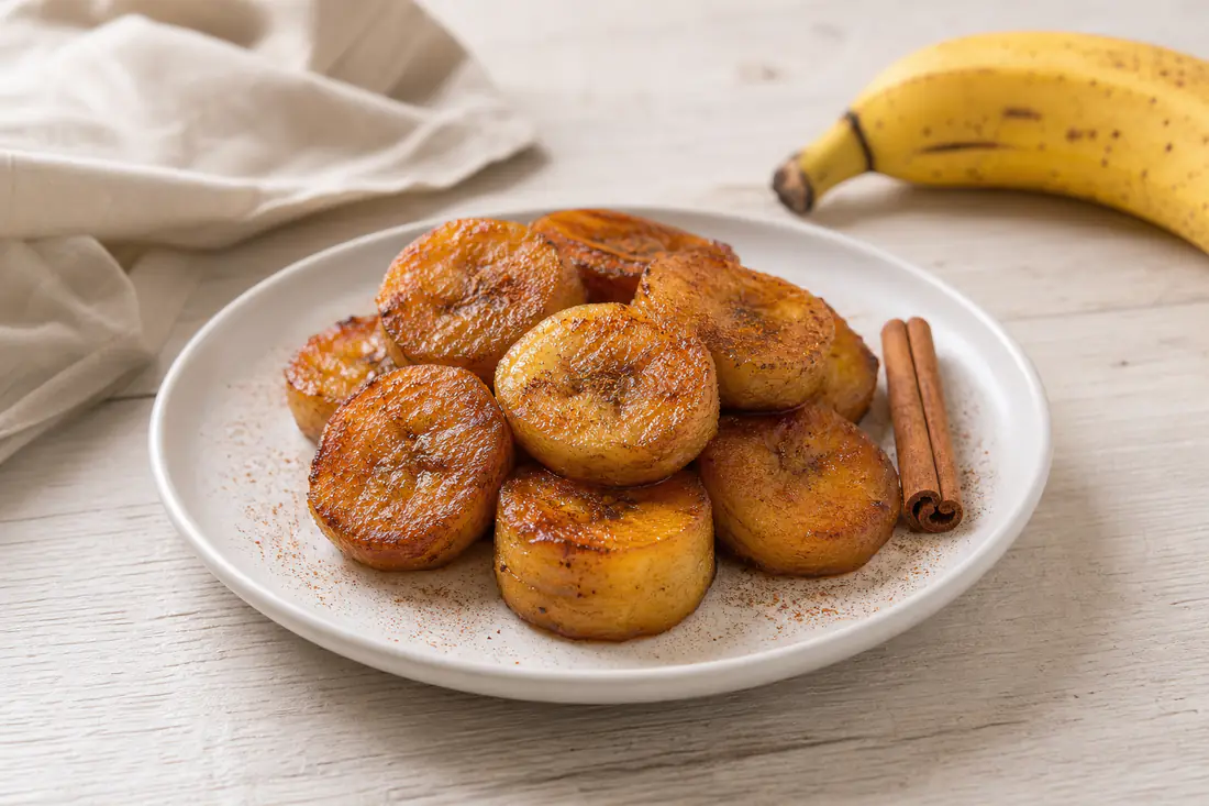 Rodelas grossas de banana frita douradas e caramelizadas, polvilhadas com canela, num prato branco com uma banana inteira ao lado