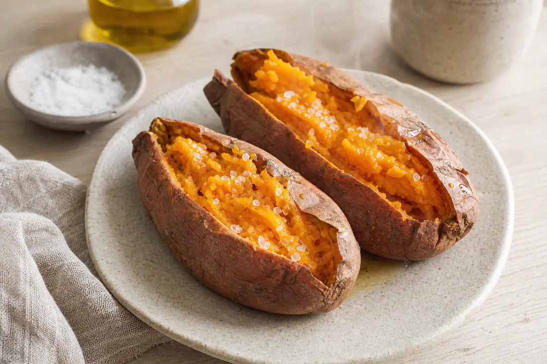 Duas batatas-doces assadas com casca, abertas ao meio, com a polpa cor de laranja a deitar vapor, com um fio de azeite e flor de sal