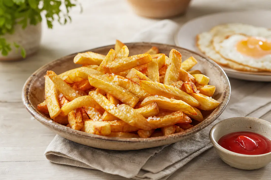 Uma travessa de batatas fritas caseiras douradas e estaladiças, polvilhadas com sal grosso, ao lado de um prato com ovo estrelado