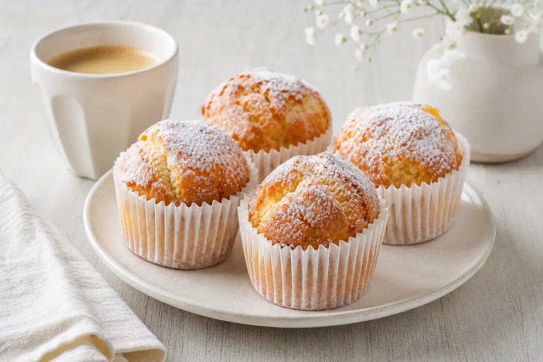 Bolinhos de arroz de pastelaria em forminhas de papel, com o topo alto, dourado e rachado, numa travessa com açúcar em pó