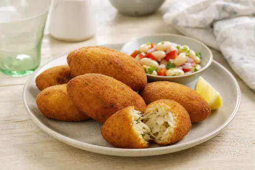 Bolinhos de bacalhau ovais e dourados, um deles aberto a mostrar o recheio de bacalhau e batata, com salada de feijão-frade