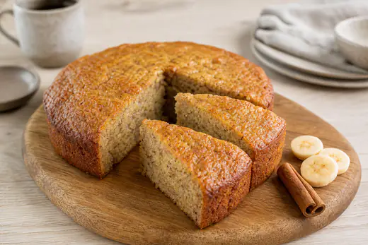 Bolo de banana redondo dourado com duas fatias cortadas, rodelas de banana e um pau de canela ao lado, sobre uma tábua de madeira