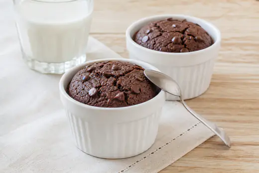 Dois mini bolos de chocolate em ramequins, com a superfície rachada e pepitas de chocolate, numa mesa com uma colher