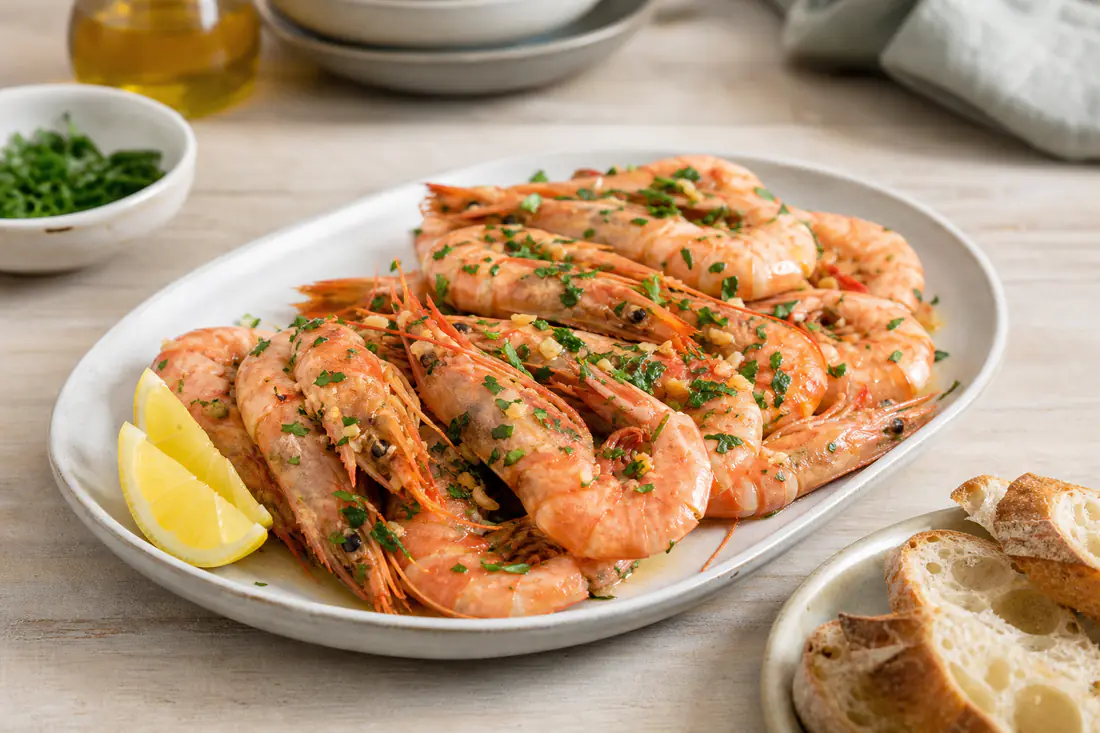 Gambas rosadas assadas com alho e salsa numa travessa, com gomos de limão e pão