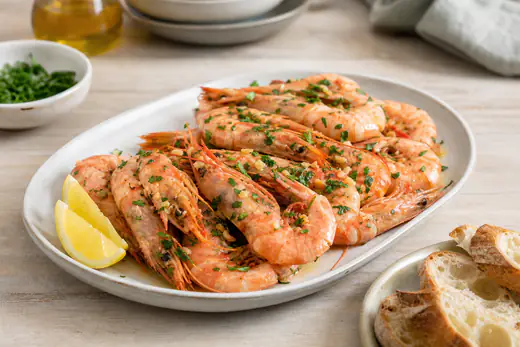 Gambas rosadas assadas com alho e salsa numa travessa, com gomos de limão e pão