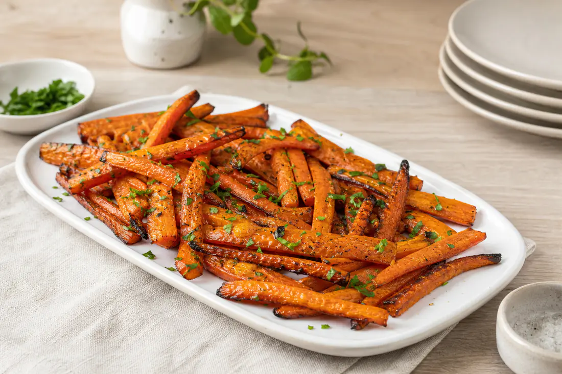 Palitos de cenoura assados, dourados nas pontas, com salsa picada, numa travessa branca