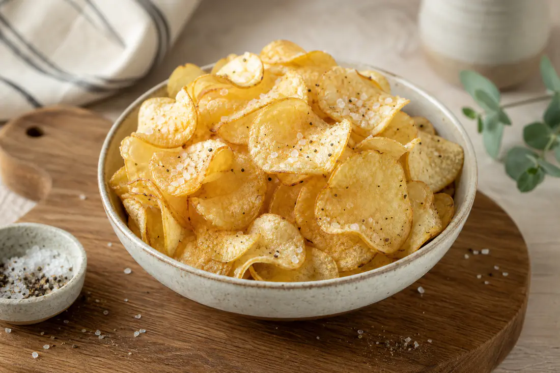 Uma taça de chips de batata caseiras, finas e douradas, com flor de sal e pimenta, sobre uma tábua de madeira