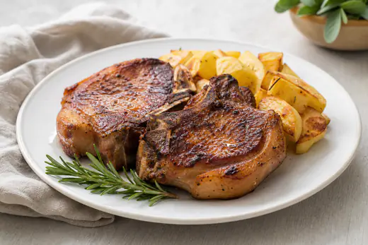 Duas costeletas de porco do cachaço com osso, bem douradas, servidas com batatas assadas e um raminho de alecrim