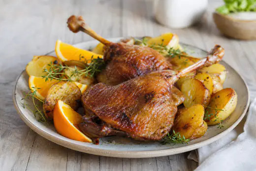 Duas pernas de pato com pele dourada e estaladiça num prato com batatas assadas e gomos de laranja