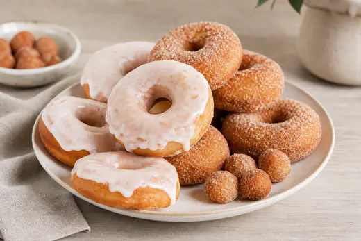 Quatro donuts dourados com cobertura de açúcar branca e outros com açúcar e canela, empilhados num prato branco, com buracos de donut ao lado