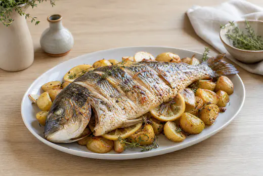 Dourada inteira assada com a pele dourada, fatias de limão e ramos de tomilho no prato, com batatas