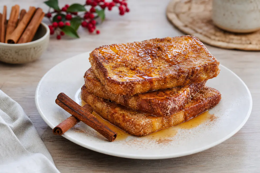 Pilha de fatias douradas de pão de forma, polvilhadas de açúcar e canela e regadas com calda, num prato branco com paus de canela ao lado