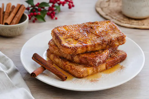 Pilha de fatias douradas de pão de forma, polvilhadas de açúcar e canela e regadas com calda, num prato branco com paus de canela ao lado