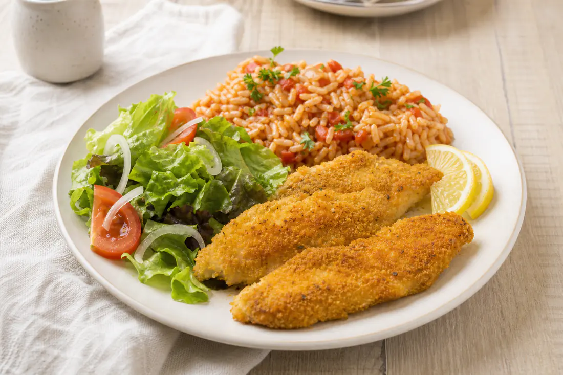 Filetes de pescada panados e dourados, servidos com arroz de tomate, salada e gomos de limão