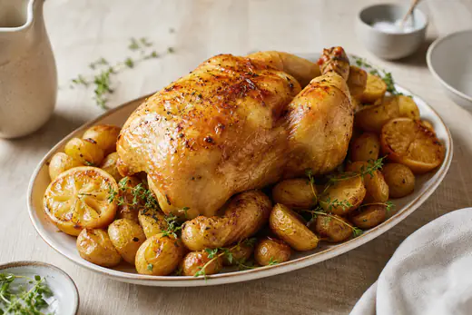 Frango inteiro assado com a pele dourada e crocante, numa travessa com batatas e raminhos de tomilho