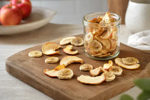 Fatias finas de maçã e rodelas de banana desidratadas, douradas e enrugadas, espalhadas numa tábua de madeira