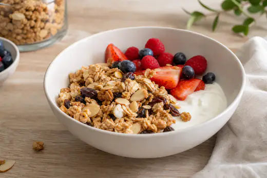Granola caseira com flocos de aveia dourados, amêndoas laminadas e passas numa taça branca com iogurte e fruta fresca