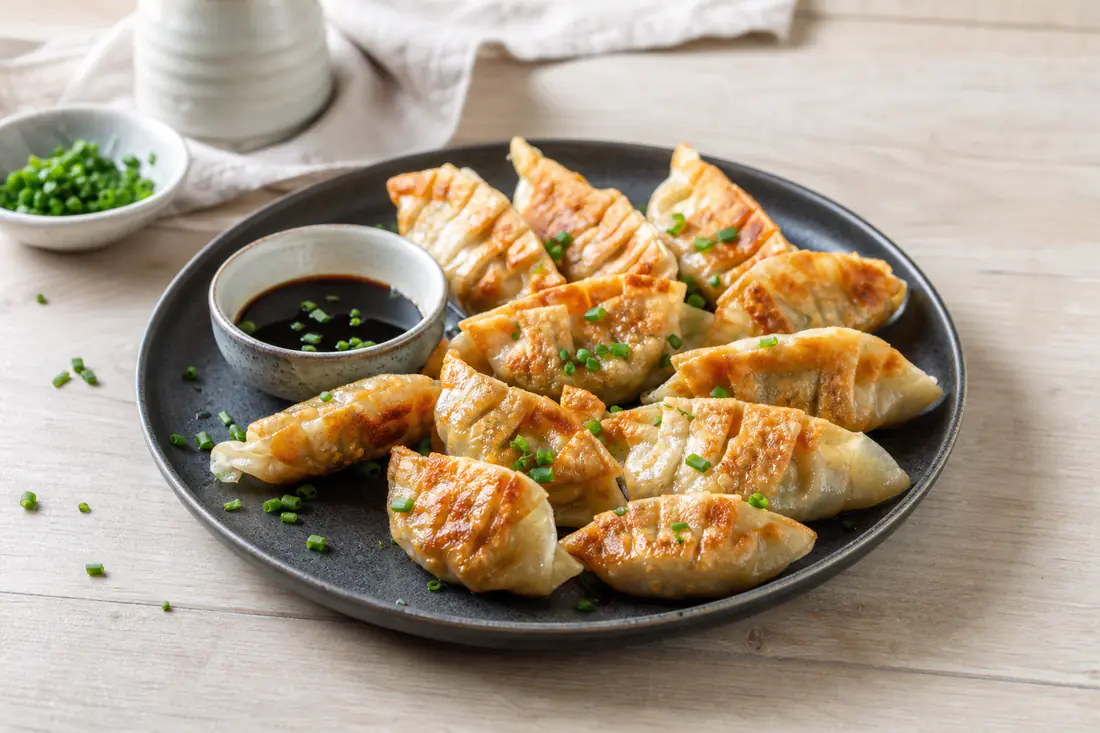 Gyozas douradas em meia-lua, com pregas na massa, num prato preto com molho de soja e cebolinho picado