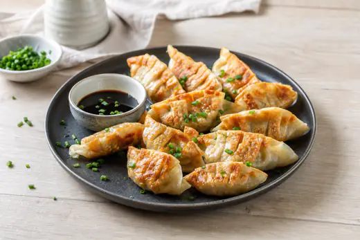 Gyozas douradas em meia-lua, com pregas na massa, num prato preto com molho de soja e cebolinho picado