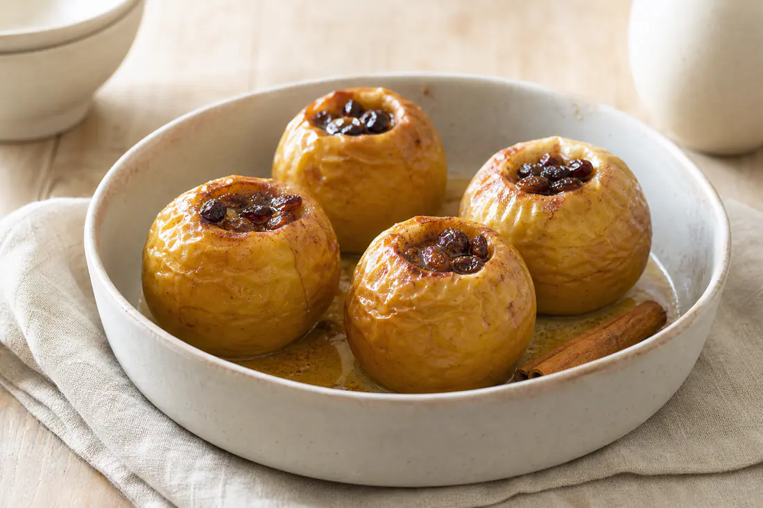 Quatro maçãs reineta assadas inteiras, com a pele enrugada e dourada e o recheio de mel, canela e passas, num prato fundo com um pau de canela