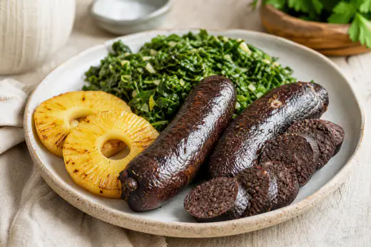 Duas morcelas assadas cortadas em rodelas grossas, com a pele escura e estaladiça, servidas com fatias de ananás e couve salteada