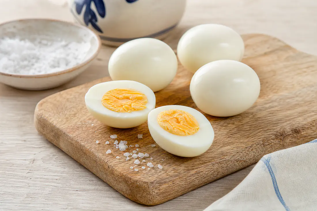 Ovos cozidos cortados ao meio com a gema amarela e cremosa, ao lado de ovos inteiros descascados numa tábua de madeira