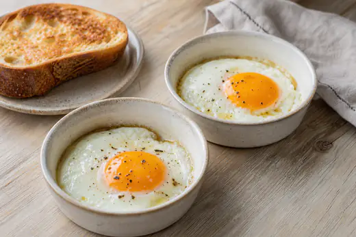 Dois ovos estrelados com a clara branca e a gema mole e brilhante, numa forma de cerâmica, com uma fatia de pão tostado ao lado