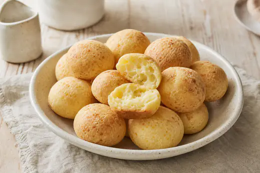 Cesto com pães de queijo redondos e dourados, um deles partido ao meio a mostrar o miolo elástico e queijoso