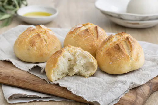 Quatro pãezinhos caseiros redondos com crosta dourada, um deles cortado ao meio para mostrar o miolo macio, numa tábua de madeira