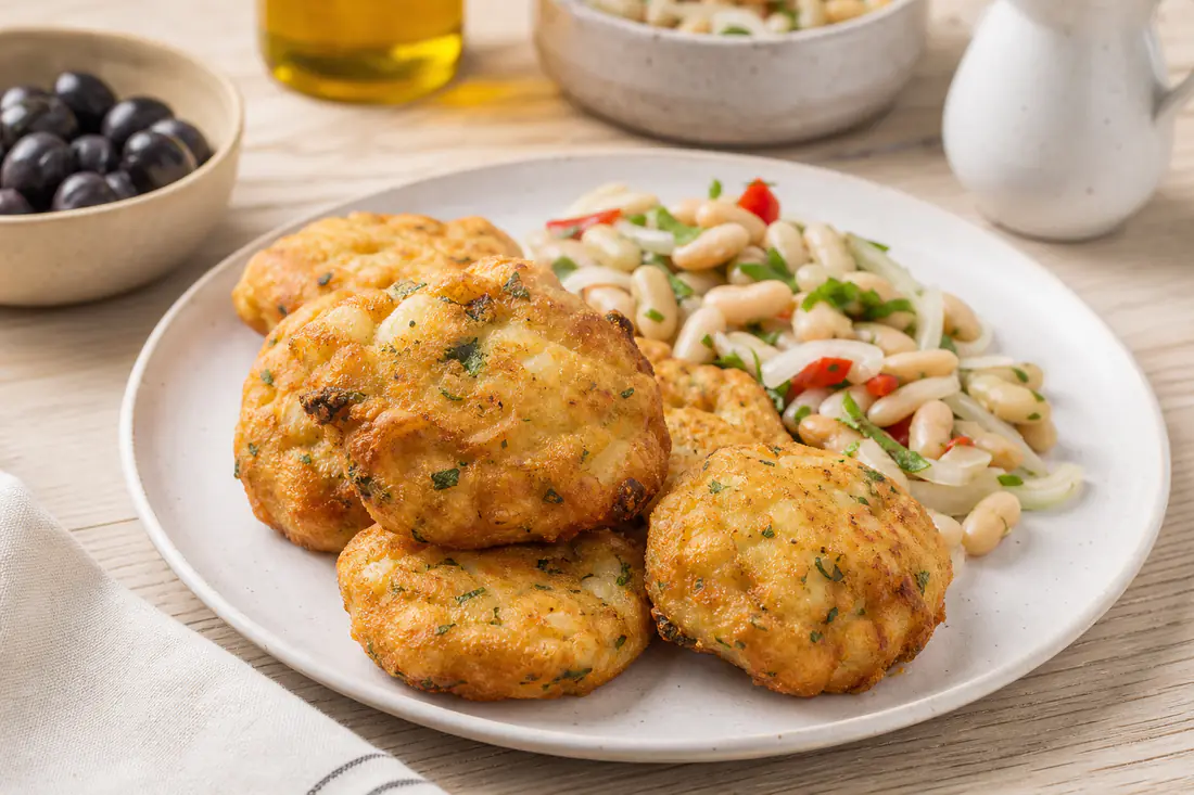 Pataniscas de bacalhau douradas, com lascas de bacalhau e salsa visíveis, num prato branco com salada de feijão-frade