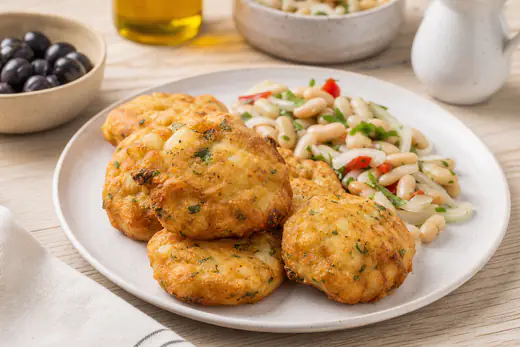 Pataniscas de bacalhau douradas, com lascas de bacalhau e salsa visíveis, num prato branco com salada de feijão-frade