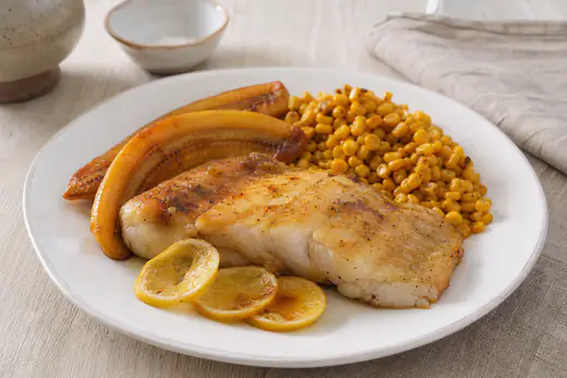 Filetes de peixe-espada preto dourados num prato, com duas bananas assadas, rodelas de limão e milho em grão
