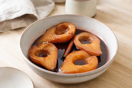 Metades de pera assada, douradas e brilhantes, numa forma com calda escura de vinho do Porto e um pau de canela, sobre uma mesa de madeira