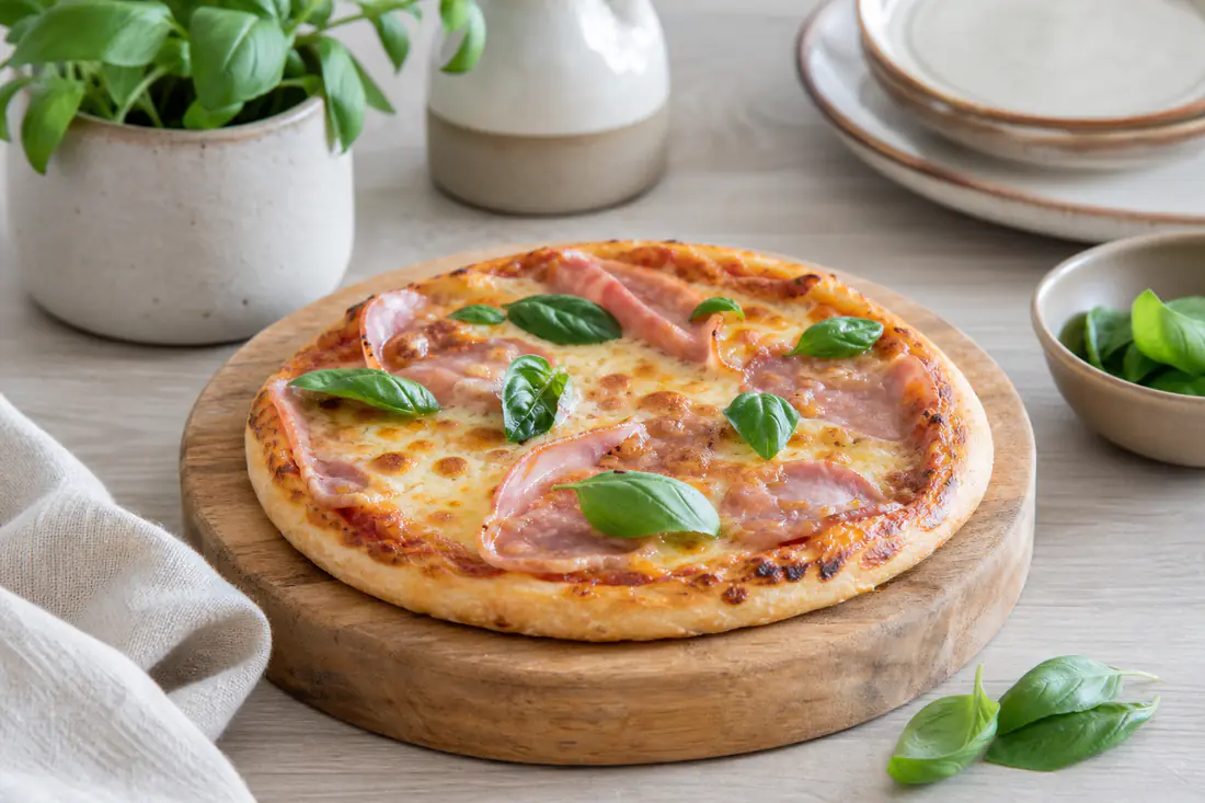 Pizza individual redonda com queijo derretido, rodelas de fiambre e manjericão fresco, com a base dourada, sobre uma tábua de madeira