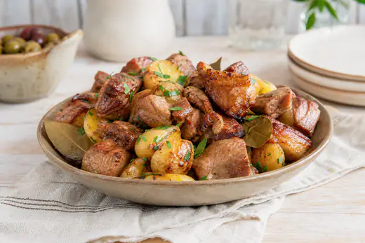 Cubos de carne de porco assada dourados, de cachaço, numa travessa com batatas assadas, folha de louro e salsa picada