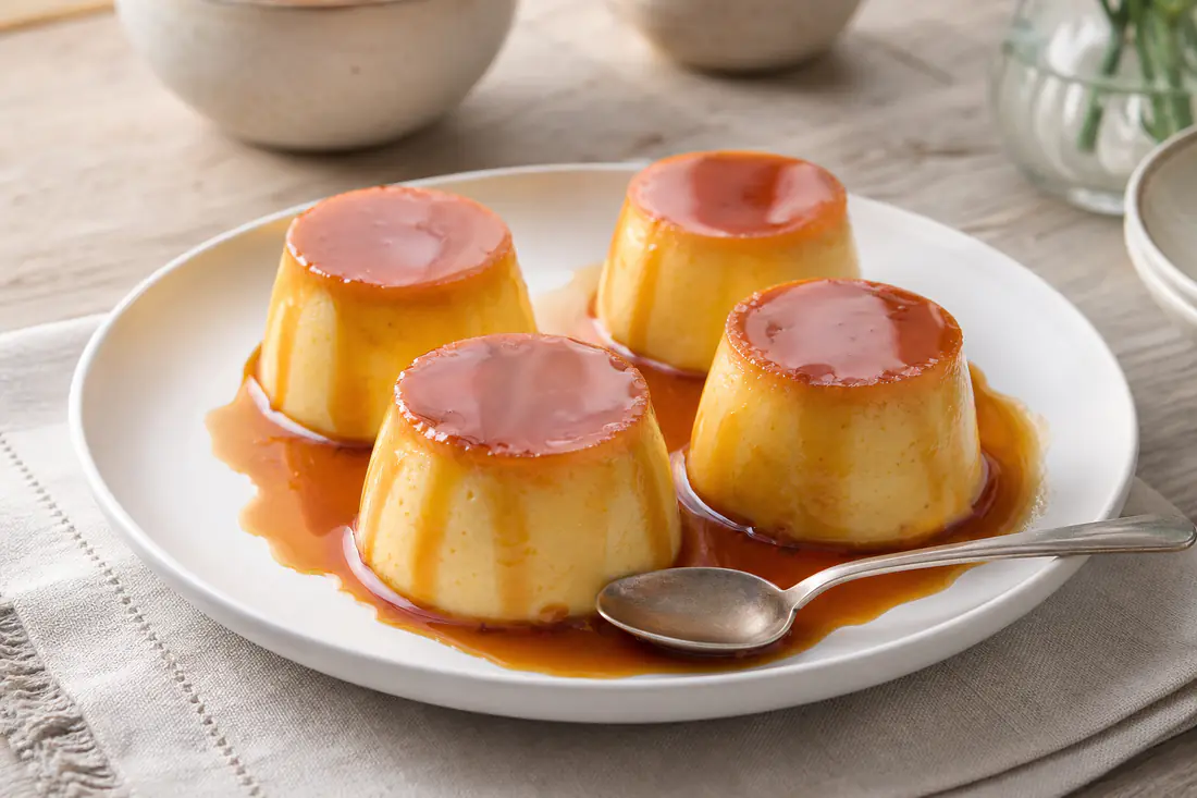 Quatro pudins individuais desenformados, com caramelo dourado a escorrer pelos lados, num prato de servir