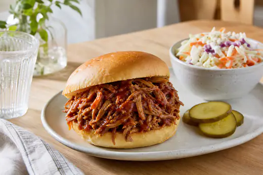 Carne de porco desfiada com molho barbecue num pão redondo, ao lado de salada de couve e pickles