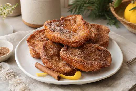 Rabanadas douradas polvilhadas com açúcar e canela, empilhadas num prato branco, com uma casca de limão e um pau de canela ao lado