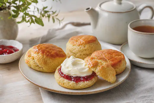 Quatro scones dourados, um deles partido ao meio, com doce de morango e natas batidas num prato, ao lado de um bule de chá