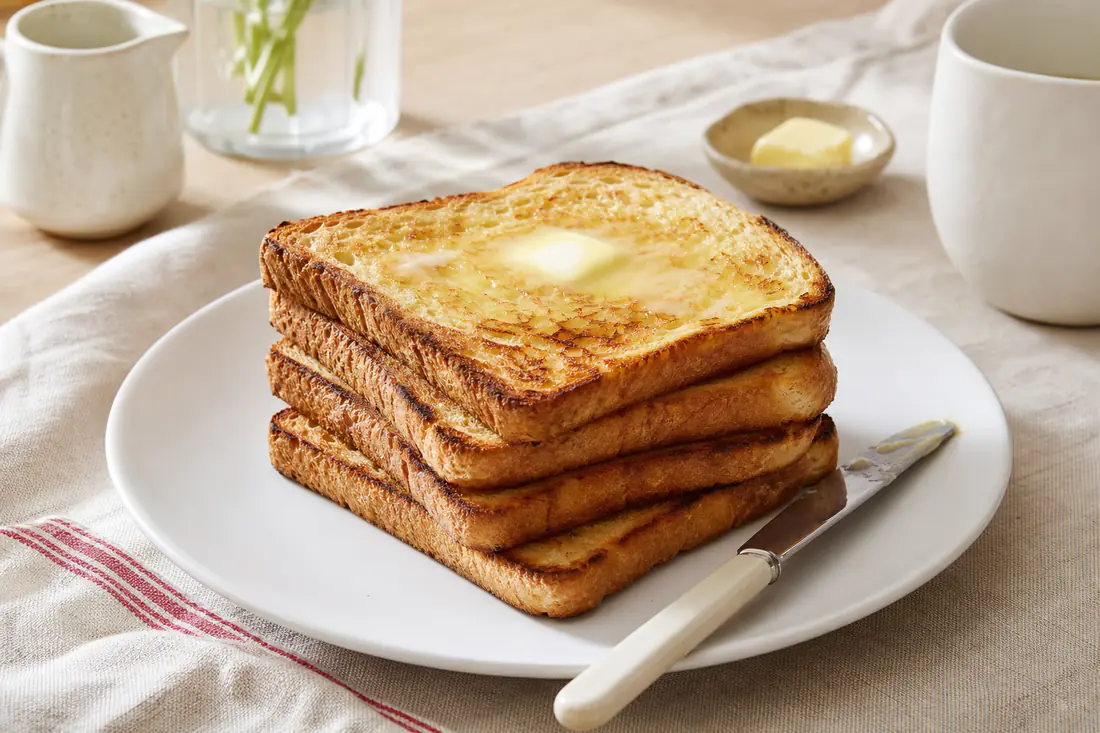 Quatro torradas de pão de forma douradas e crocantes, empilhadas num prato, com manteiga a derreter e uma faca ao lado