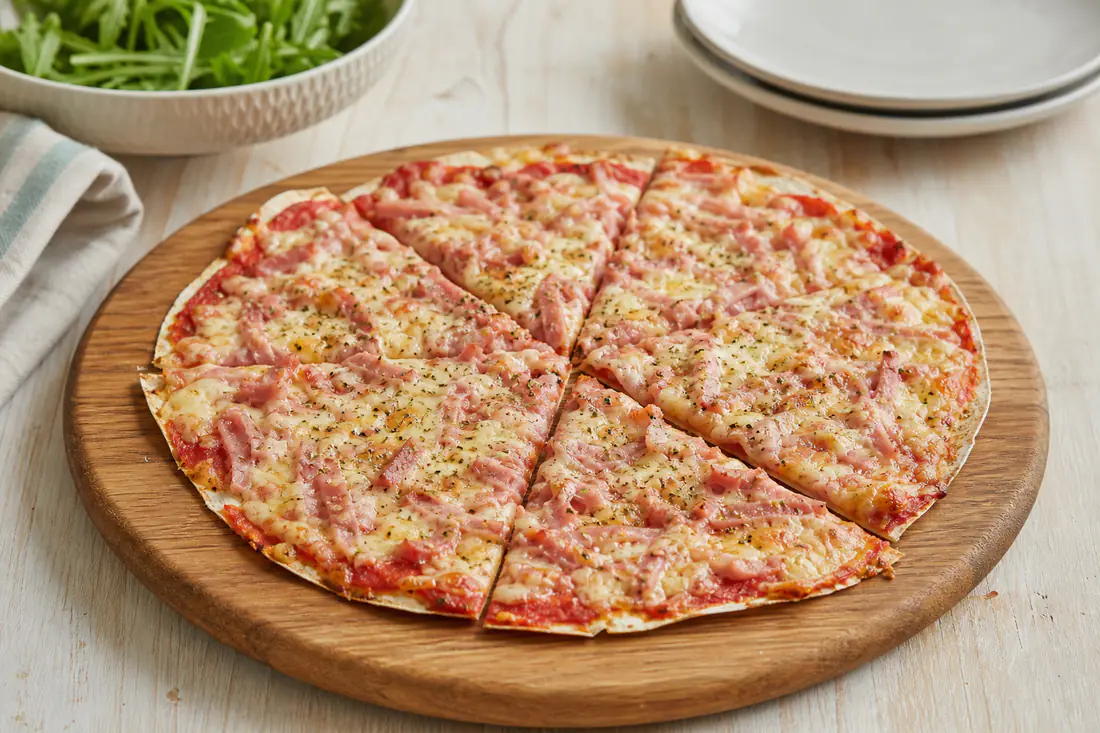 Pizza feita com um wrap fino, com molho de tomate, queijo derretido, tiras de fiambre e orégãos, cortada em fatias numa tábua